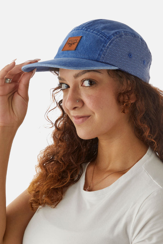 A woman with vibrant red curly hair wears a white shirt and a denim-styled cap, facing left. Her right hand touches the visor. The blue cap features a brown sewed-on patch with the iconic Cat School symbol from The Witcher universe on the front side.