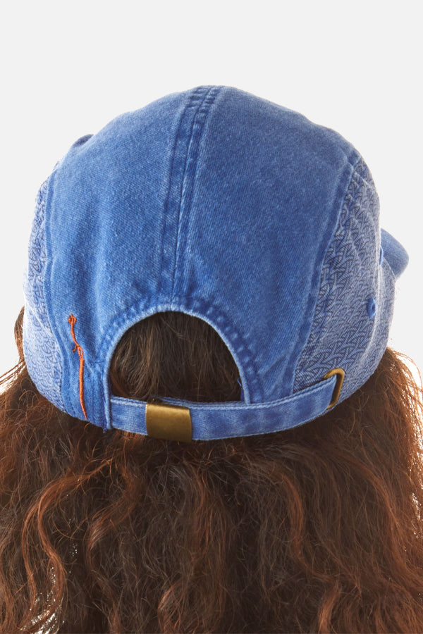 The back of a person's head is visible, adorned with a blue denim-styled cap. Dark red curly hair emerges from under the cap, with an adjustable strap on top.