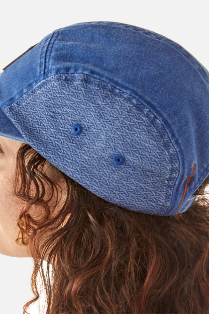 The top profile of a person is visible, showcasing the side of a blue denim cap with a pattern. Dark red curly hair is visible under the cap, peeking out from beneath.