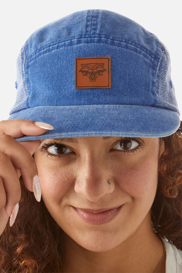 We see a woman with brown eyes, curly red hair, and a nose piercing, wearing a blue denim-styled cap known as 'The Witcher Cat School Hat.' She is touching the front visor, presenting a sewed-on patch featuring the Cat School symbol from The Witcher universe.