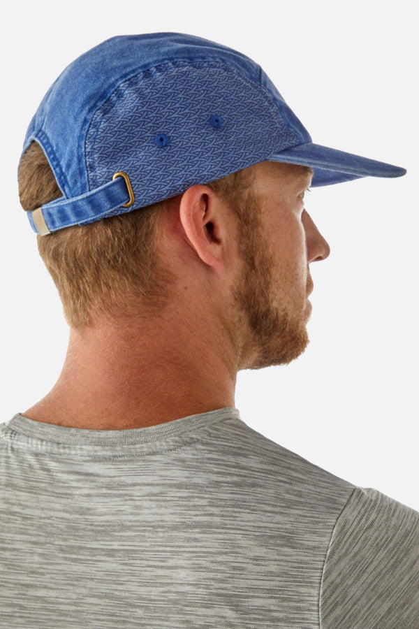 We see an upper profile of a male model from behind. The man faces the reght side and  has a light blond beard and short blond hair, wearing a white striped shirt. He sports a blue denim-styled cap with a pattern on the left side, and at the back of his head, we can see the adjustable strap.